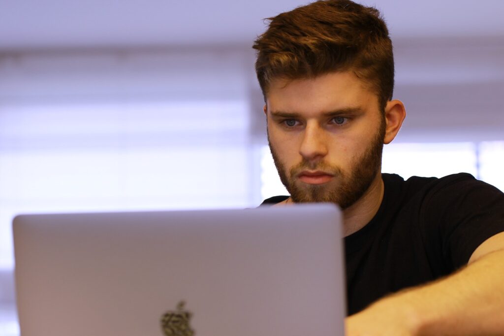 Na imagem, um rapaz observa atentamente a tela de seu computador, enquanto trabalha em um projeto de Engenharia de Produção. A cena reflete o foco necessário para desenvolver soluções baseadas em dados e otimização de processos, aplicando conceitos aprendidos durante sua formação acadêmica. Ele representa estudantes e profissionais que utilizam a tecnologia para criar inovações no ambiente industrial.